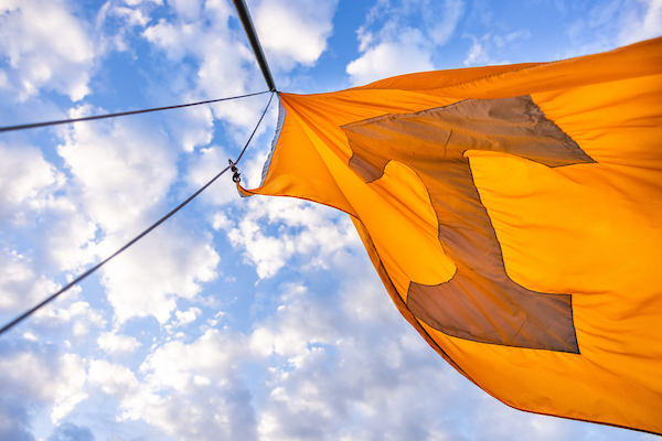 A an orange Power "T" flag, for the University of Tennessee, waves against a bright blue, cloudy sky.